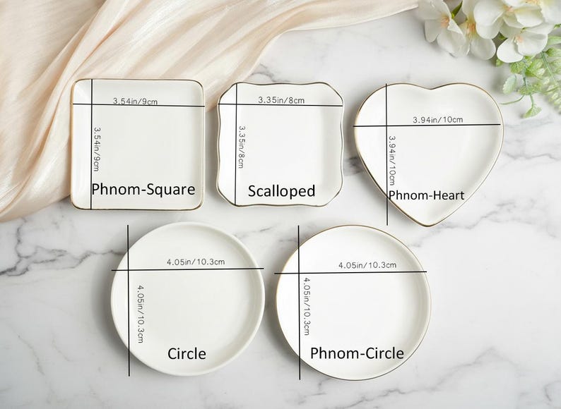 May include: A collection of white ceramic plates in various shapes: square, scalloped, heart, and two circles. Each plate has a gold rim and measurements in inches and centimeters. The square plate is 3.54 inches (9cm) and the heart plate is 3.94 inches (10cm).