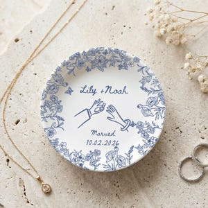 May include: A white ceramic ring dish with a blue floral border and the text "Lily + Noah Married 10.02.2026". The dish features a design of two hands exchanging rings. A gold necklace and two rings are also present.
