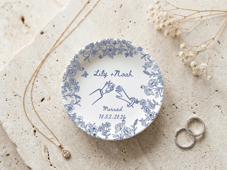 May include: A white ceramic ring dish with a blue floral border and the text "Lily + Noah Married 10.02.2026". The dish features a design of two hands exchanging rings. A gold necklace and two rings are also present.