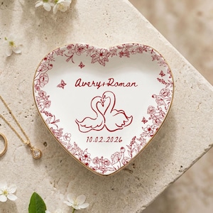 May include: Heart-shaped ceramic trinket dish with a floral border and gold trim. The dish features a design of two swans and the names "Avery + Roman" with the date "10.02.2026". A gold necklace and ring are also visible.