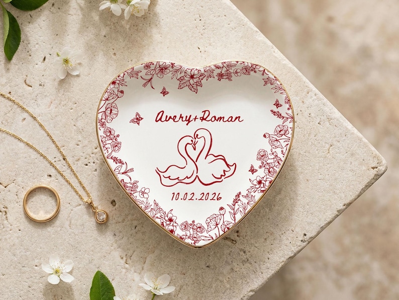 May include: Heart-shaped ceramic trinket dish with a floral border and gold trim. The dish features a design of two swans and the names "Avery + Roman" with the date "10.02.2026". A gold necklace and ring are also visible.