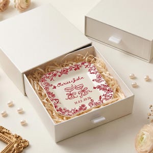 May include: A square, off-white ceramic plate with a red floral border and matching text, presented in a white gift box. The plate is inscribed with "Clarise & John" and a date. The box is filled with straw.