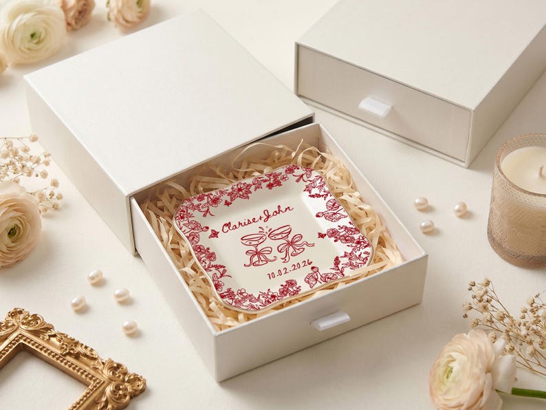 May include: A square, off-white ceramic plate with a red floral border and matching text, presented in a white gift box. The plate is inscribed with "Clarise & John" and a date. The box is filled with straw.