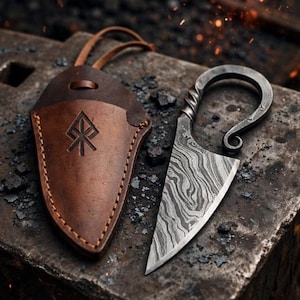 Op de afbeelding: Een klein mes met vaststaand lemmet van patroonrijk Damascusstaal en een gekruld metalen handvat, vergezeld door een bruine lederen schede met een gestempeld runensymbool en een leren koord, rustend op een metalen oppervlak.