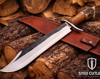 May include: A large, handmade hunting knife with a polished steel blade and a brown leather handle. The knife rests on a brown leather sheath, set against a backdrop of stacked firewood. The knife has a decorative handle and is a product of Syed Cutlery.