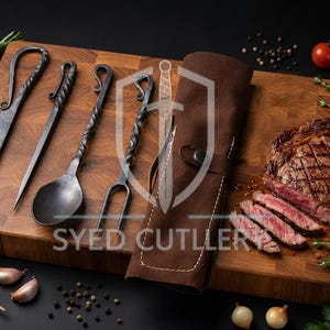 May include: A set of four hand-forged iron utensils including a knife, a skewer, a spoon, and a two-pronged fork, each featuring a twisted handle. The set is accompanied by a brown leather roll-up case with the text SYED CUTLERY and a shield logo.