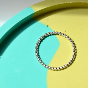 Könnte beinhalten: Ein silberner Ring mit einem Perlendesign. Der Ring befindet sich auf einem zweifarbigen Tablett mit türkisfarbenem und gelbem Farbschema. Die kreisförmige Form des Rings wird durch die kontrastierenden Farben hervorgehoben.
