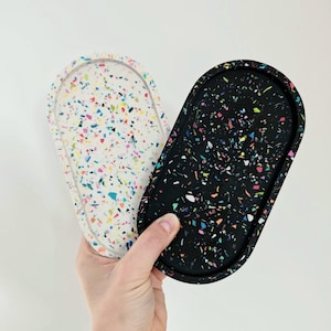 Op de afbeelding: Twee ovale trays met terrazzo-stijl ontwerpen. Eén tray is wit met kleurrijke vlekken, de andere zwart met vergelijkbare accenten. De trays worden tegen een witte achtergrond gehouden.