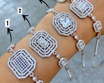 Op de afbeelding: Vier zilveren armbanden met ingewikkelde, geometrische ontwerpen worden getoond. Elke armband heeft een centraal, versierd vierkant of rechthoekig element, omgeven door kleinere, sprankelende accenten. De armbanden zijn verbonden door fijne kettingen en hebben een bungelend, decoratief uiteinde.