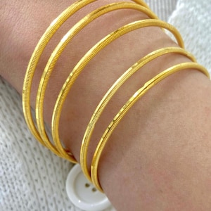 Pode incluir: Um conjunto de cinco pulseiras circulares finas de ouro com uma superfície texturizada e canelada. As pulseiras são usadas juntas, criando uma aparência empilhada. Cada pulseira apresenta um acabamento amarelo metálico com sulcos finos.
