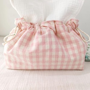 Puede incluir: Una funda para caja de pañuelos de tela de cuadros rosas y blancos con cierre de cordón. El tejido blanco es visible en la parte superior. A la derecha hay un pequeño paño plegado con estampado floral.