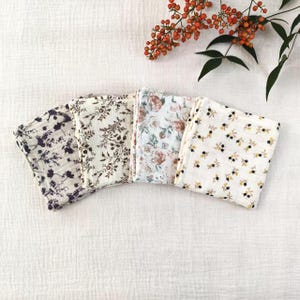 May include: Five folded floral patterned cloths in various designs. The cloths feature patterns of purple, brown, and yellow flowers on a white background. A sprig of orange berries and green leaves is in the upper right corner.