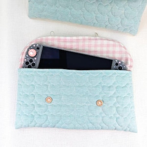 May include: A light blue quilted fabric pouch with a heart pattern containing a handheld gaming console. The pouch features a pink and white gingham interior lining and two small wooden buttons on the front flap for closure.