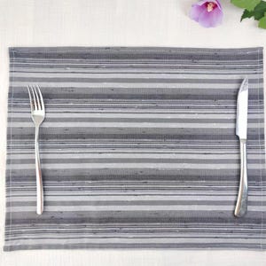 May include: A rectangular placemat featuring a horizontal striped pattern in various shades of grey, charcoal, and white. A silver-toned metal fork is positioned on the left side, and a silver-toned metal dinner knife is on the right side.