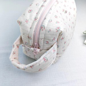 May include: A cream-colored quilted pouch featuring a delicate pink and green floral pattern with thin vertical stripes. The bag includes a matching fabric handle on the side and a light pink zipper closure with a small flower-shaped zipper pull.