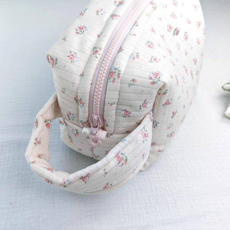 May include: A cream-colored quilted pouch featuring a delicate pink and green floral pattern with thin vertical stripes. The bag includes a matching fabric handle on the side and a light pink zipper closure with a small flower-shaped zipper pull.