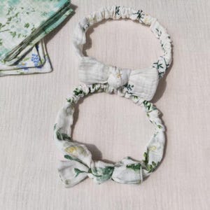 Puede incluir: Dos diademas blancas con lazos. Las diademas están adornadas con un delicado estampado floral en tonos verdes y amarillos. La tela parece suave y ligera, adecuada para el clima cálido.