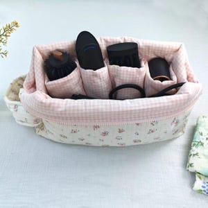 Può includere: Un organizer in tessuto trapuntato con esterno color crema con una piccola stampa floreale rosa e verde e un interno a quadretti vichy rosa chiaro e bianco. L'organizer presenta diversi scomparti interni contenenti vari strumenti per lo styling neri.