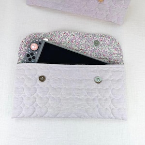 May include: A light lavender quilted fabric pouch featuring a heart-shaped stitching pattern and two silver metal snap buttons. The interior flap is lined with a floral print fabric, and a grey handheld gaming console is tucked inside the pouch.