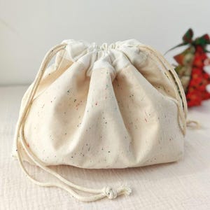 Könnte beinhalten: Ein cremefarbener Kordelzugbeutel mit bunten Sprenkeln. Die Tasche ist aus einem weichen Stoff gefertigt und hat einen passenden Kordelzugverschluss. Ein kleiner, dekorativer Weihnachtsbaum befindet sich im Hintergrund.