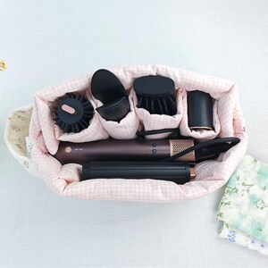 May include: A soft, quilted storage bag with a light pink and white gingham pattern containing a dark brown hair styling tool with multiple black attachments, including a round brush, a concentrator nozzle, and a cylindrical curling barrel.