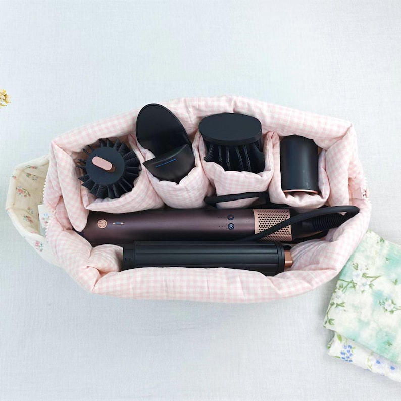 May include: A soft, quilted storage bag with a light pink and white gingham pattern containing a dark brown hair styling tool with multiple black attachments, including a round brush, a concentrator nozzle, and a cylindrical curling barrel.