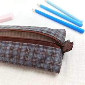 May include: A quilted pencil case featuring a blue and brown plaid pattern with a brown zipper closure. The interior is lined with a light-colored gingham fabric. Three blue pencils and a small pink eraser are positioned near the case.