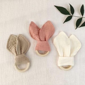 May include: Three baby teething toys in soft, muted colors. Each toy features a wooden ring base with fabric bunny ears in beige, dusty rose, and cream. The toys are made of a textured, crinkled fabric.