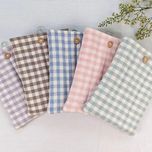 May include: Five rectangular fabric pouches in pastel colors: lavender, brown, blue, pink, and mint green. Each pouch features a gingham pattern and a wooden button closure. The pouches are arranged on a white surface.