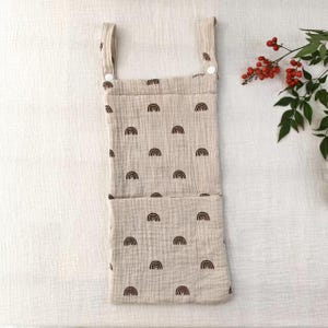 May include: A beige hanging storage organizer made of textured muslin fabric featuring a repeating pattern of small brown rainbows. The organizer has two top straps with white snap buttons for attachment and a large front pocket.