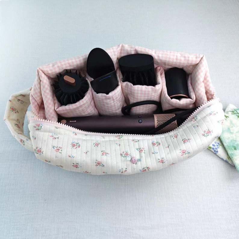 May include: A quilted fabric storage bag with a cream base and small pink floral print, featuring a zipper closure. The interior is lined with pink and white gingham fabric and contains several black hair styling tools and attachments.