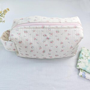 May include: A cream-colored quilted pouch featuring a repeating pattern of small pink and blue flowers. The pouch has a light pink zipper closure along the top, a side handle, and a textured fabric surface with vertical stitching throughout the exterior.