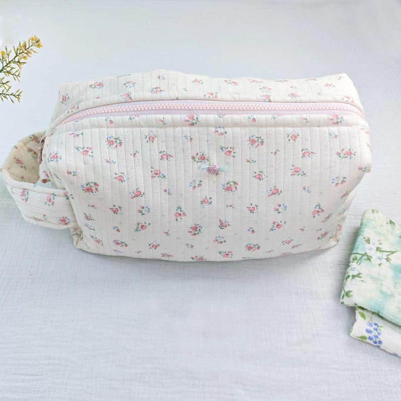 May include: A cream-colored quilted pouch featuring a repeating pattern of small pink and blue flowers. The pouch has a light pink zipper closure along the top, a side handle, and a textured fabric surface with vertical stitching throughout the exterior.
