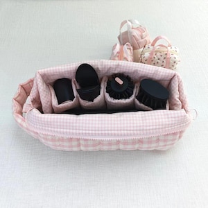 May include: A large, rectangular, quilted makeup bag with a light pink and white gingham pattern, featuring a zippered top and internal dividers holding four black cylindrical hair styling tool attachments. Two smaller matching bags sit behind.