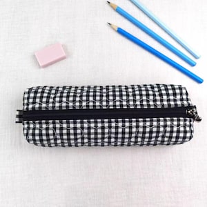 May include: A rectangular pencil case featuring a black and white gingham pattern with quilted heart-shaped stitching. The case has a black zip closure along the top. A light pink rectangular eraser and three blue pencils are positioned nearby.