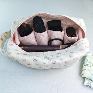 May include: A quilted fabric storage bag featuring a cream background with a small pink and blue floral print. The interior is lined with a pink and white gingham fabric and contains multiple compartments holding various hair styling tools.