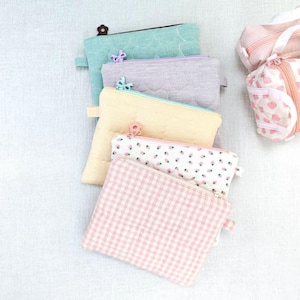 May include: A collection of five rectangular zipped pouches in various colours and patterns, including solid teal, lavender, and yellow, a white pouch with a small strawberry print, and a pink and white gingham check pouch with a flower zip pull.