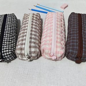 May include: Four rectangular quilted pencil cases with zippered closures. The first is black and white gingham, the second is beige and white gingham, the third is pink and white heart-patterned, and the fourth is a blue and brown plaid.