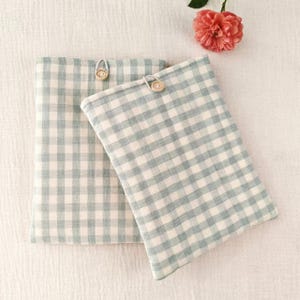 May include: Two rectangular, light blue and white gingham fabric cases with wooden button closures. The cases are on a white surface, with a pink flower. The cases appear to be for books or tablets. The gingham pattern is a classic design.