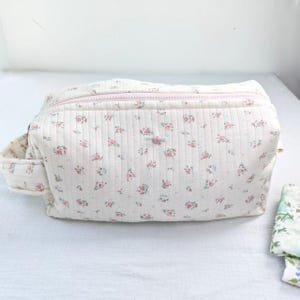 May include: A rectangular quilted pouch featuring a cream-colored fabric with a repeating pattern of small pink roses and tiny blue leaves. The bag includes a light pink zipper closure along the top and a small fabric handle on the left side.