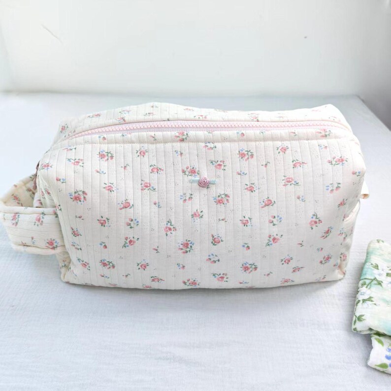 May include: A rectangular quilted pouch featuring a cream-colored fabric with a repeating pattern of small pink roses and tiny blue leaves. The bag includes a light pink zipper closure along the top and a small fabric handle on the left side.