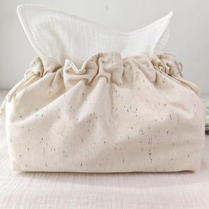 May include: A beige fabric tissue box cover with a drawstring closure. The cover has small flecks of red, blue, and green. White tissues are visible at the top. A folded cloth with a floral pattern is in the background.