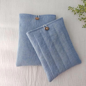 May include: Two light blue fabric cases with wooden button closures. One case is quilted with vertical lines. The cases are on a white textured surface, with a sprig of greenery in the upper right corner.