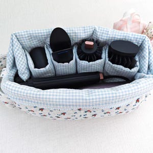 May include: A fabric storage organizer with a white and blue gingham interior and a floral exterior pattern. The organizer features multiple vertical compartments holding black hair styling tools, including a flat iron and various brush attachments.