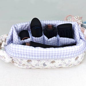 May include: A quilted fabric storage organizer with a light purple and white gingham interior lining and a floral patterned exterior. The organizer features multiple internal compartments holding various black hair styling tools and attachments.