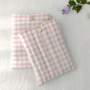 May include: Two rectangular, pink and white gingham fabric pouches with wooden button closures. The pouches are made of a soft, textured material. The background is a white, textured fabric. A sprig of greenery with a purple flower is in the upper right corner.