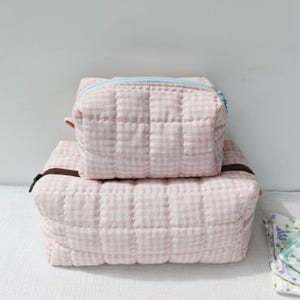 Op de afbeelding: Twee rechthoekige, gewatteerde etuis met een lichtroze en wit gingham-patroon. Het kleinere bovenste etui heeft een lichtblauwe rits, terwijl het grotere onderste etui is voorzien van een donkerbruine rits.