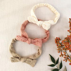 May include: Three baby headbands in cream, dusty rose, and tan, each with a knotted bow. The headbands are arranged on a white, textured surface, with a sprig of orange berries and green leaves in the background.