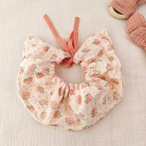 May include: A baby bib featuring a pink rose pattern on a cream backdrop. The bib has a gathered neckline and a peach-coloured tie closure. The bib is made of soft, lightweight fabric.
