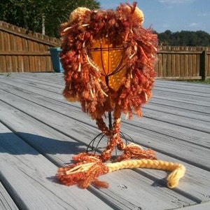 May include: A handmade lion hat with a mane of orange, brown, and yellow yarn. The hat has two small ears and a braided tail. The hat is displayed on a wire stand on a wooden deck.